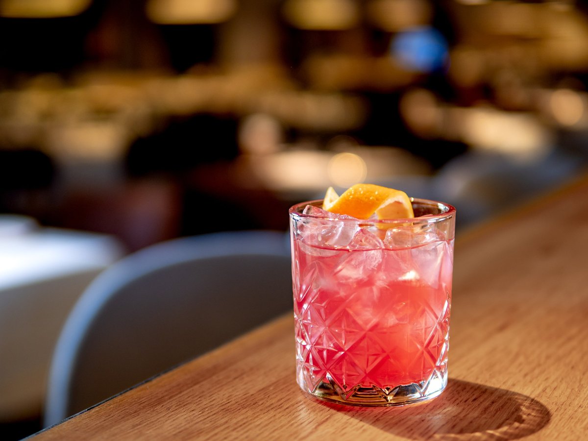 A transparent pink aperol and gin cocktail with a curled orange zest spiral, set on the bar counter at Sip Sip