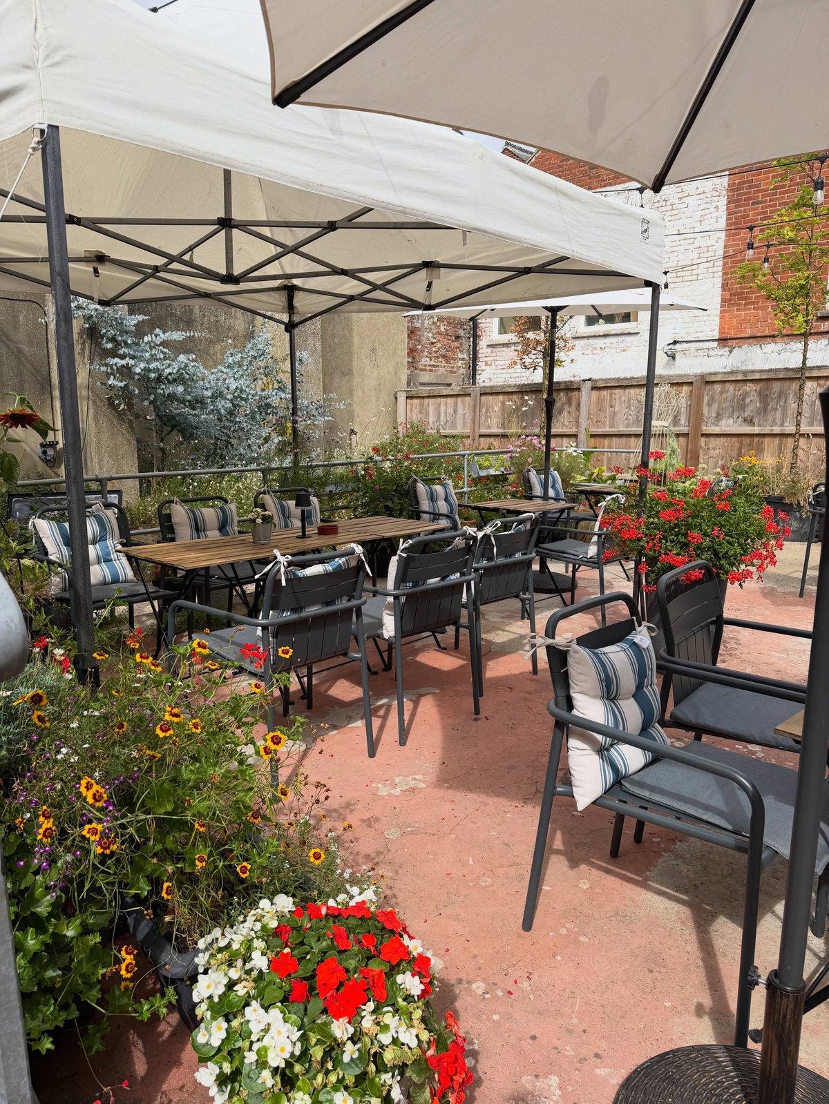 Outdoor patio dining area with metal table, striped chairs, umbrellas, and potted red and white flowers